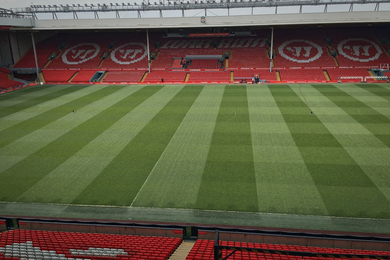 Empty Anfield pitch with striped grass in front of a red stand with large white logos in the seats