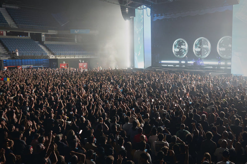 Packed arena crowd with raised arms and phone lights facing a brightly lit concert stage