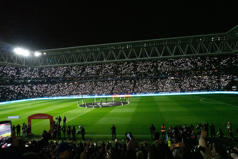 Full stadium bowl at night with both teams lined up at the centre circle