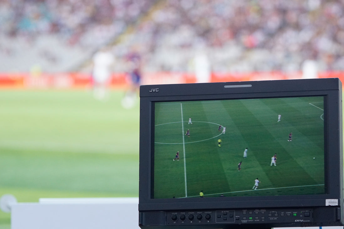 Pitchside broadcast monitor showing a live football match, with the stands blurred behind