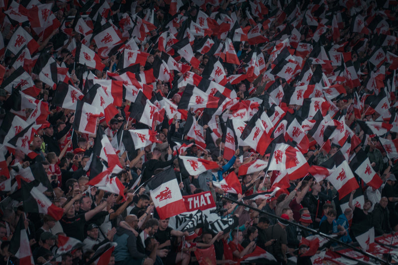 A large crowd of fans waving Manchester United flags in a stadium or public gathering, with many flags showing the red devil crest and some holding signs.