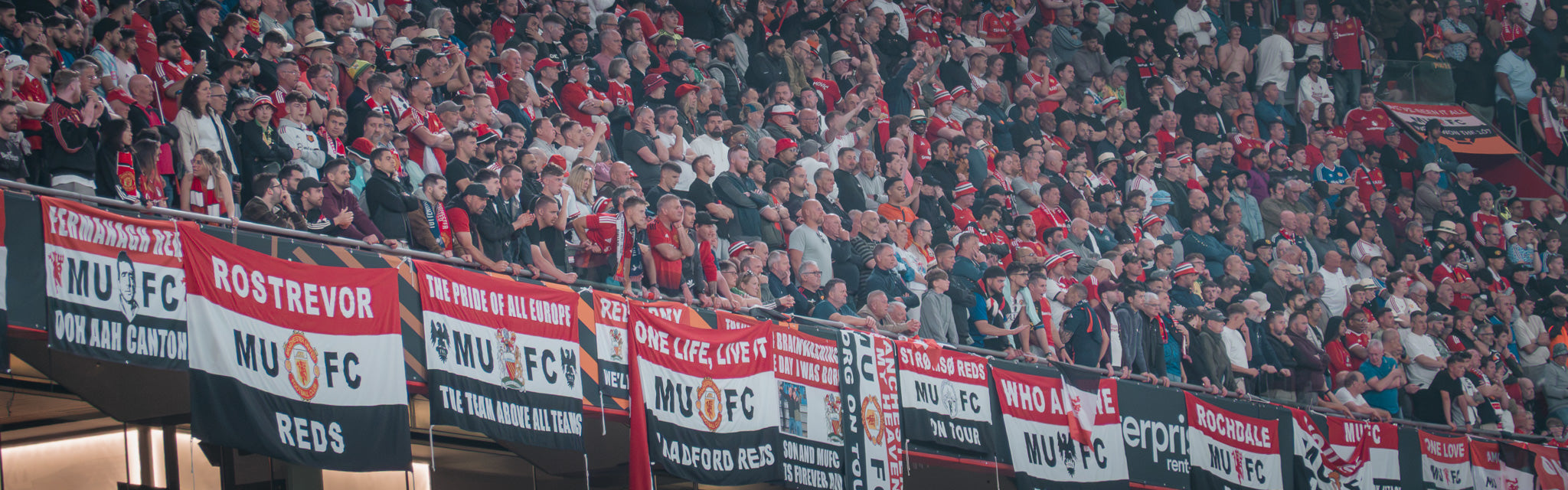 A crowded stadium crowd wearing red and white, with multiple banners reading ROSTREVOR and MUFC.