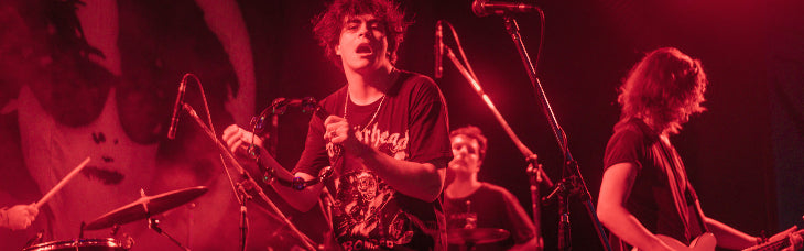 Winona Riders performing on stage under heavy red stage lighting, with a vocalist holding a microphone in the center, a drummer on the left, and a guitarist on the right.